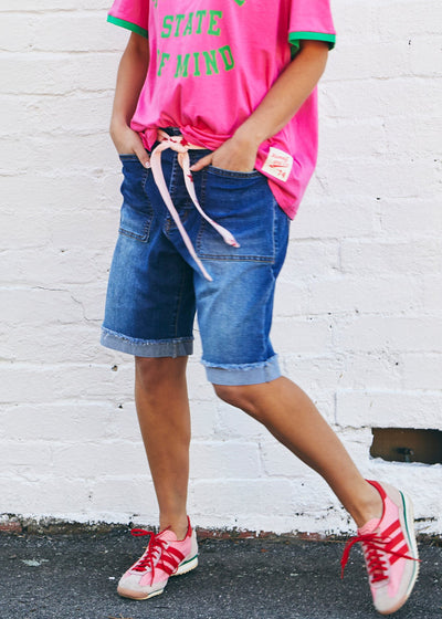 Hammill & Co A pair of longer-length darker denim stretch shorts with a relaxed fit. They have front pockets, a raw rolled hem, and sit mid-thigh. 